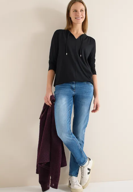 Frau in schwarzer Bluse, blauen Jeans und Sneakern hält eine dunkelrote Jacke.